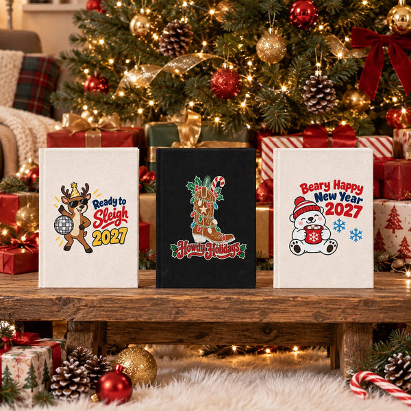 Three Christmas and New Year 2027 canvas notebooks, natural and black, with Ready to Sleigh reindeer, Howdy Holidays cowboy boot and Beary Happy New Year polar bear prints in front of a decorated Christmas tree and presents
