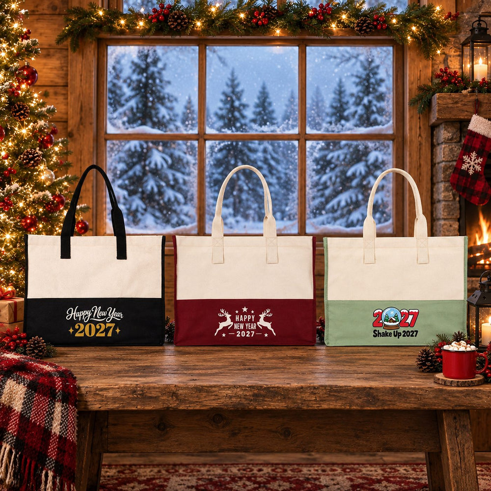 Three large two-tone canvas tote bags in black, burgundy and sage green with Happy New Year 2027 script, leaping reindeer and Shake Up 2027 snow globe prints on a wooden bench in front of a snowy cabin window at Christmas
