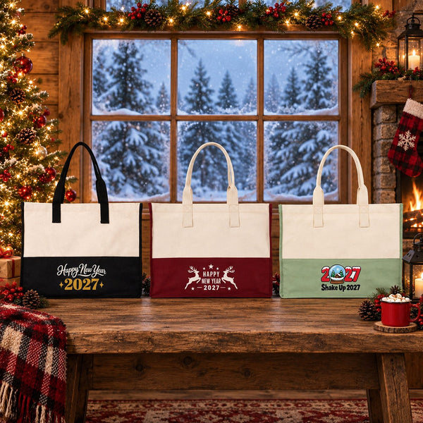 Three large two-tone canvas tote bags in black, burgundy and sage green with Happy New Year 2027 script, leaping reindeer and Shake Up 2027 snow globe prints on a wooden bench in front of a snowy cabin window at Christmas