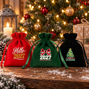 Three velvet drawstring gift pouches in red, green and black with gathered rose tops and satin ribbons, printed with Hello 2027, buffalo plaid bow 2027 and Merry Christmas tree designs, on a snowy rustic table in front of a lit Christmas tree