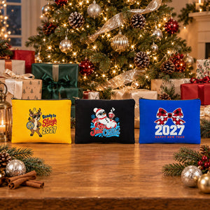 Three New Year 2027 zippered clutch bags in yellow, black and royal blue with Ready to Sleigh reindeer, Just Chillin snowman and buffalo plaid bow 2027 prints on a table in front of a decorated Christmas tree and presents