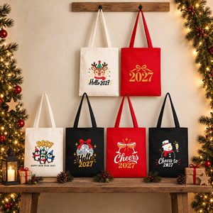 Six Christmas 2027 cotton gabardine tote bags in natural, red and black, two hanging on a wooden rail and four standing on a rustic bench, with reindeer, gold 2027, penguin, disco ball, Cheers 2027 and snowman prints
