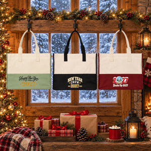Three large canvas tote bags in sage green, black and burgundy with Happy New Year 2027 script, New Year Crew 2027 and Shake Up 2027 snow globe prints hanging from iron hooks in front of a snowy cabin window at Christmas