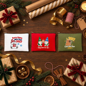 Three printed cotton clutch bags, white, red and olive green, with Beary Happy New Year polar bear, Happy Gnome Year 2027 and Capy New Year 2027 prints on a gift-wrapping table
