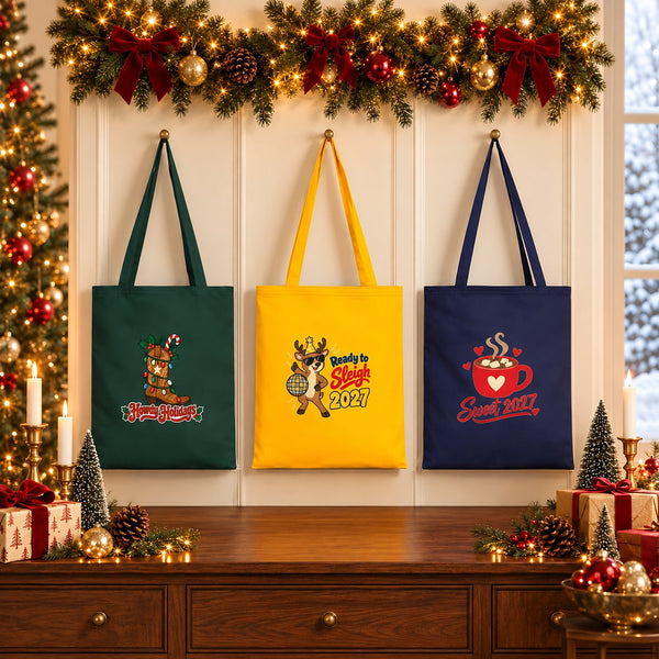 Three New Year 2027 gabardine tote bags, 14 x 16 inches, in green, yellow and navy hanging above a festive Christmas sideboard with Howdy Holidays, Ready to Sleigh 2027 and Sweet 2027 prints