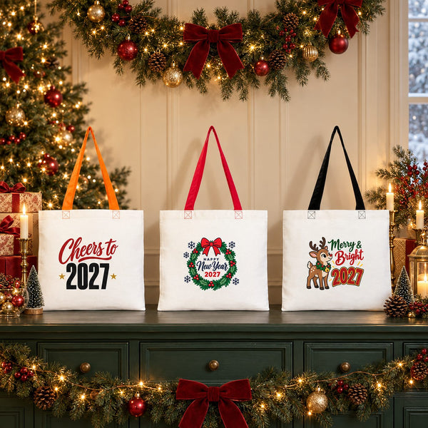Three New Year 2027 cotton gabardine tote bags, 16 x 14 inches, with orange, red and black handles standing on a festive Christmas sideboard with Cheers to 2027, wreath and reindeer Merry and Bright 2027 prints