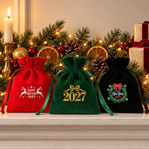 Red, green and black velvet drawstring pouches printed with Happy New Year 2027 reindeer, gold bow 2027 and holly wreath designs standing on a white fireplace mantel with a lit garland, a brass candle and wrapped gifts