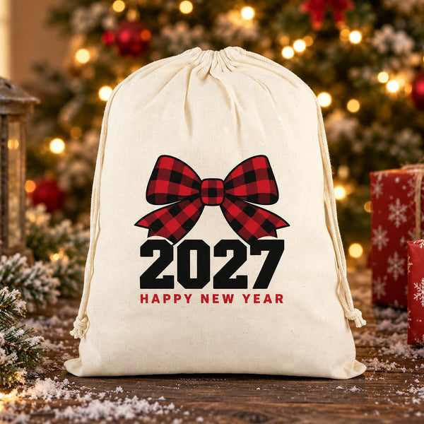 Natural cotton drawstring bag with a gathered top and hanging cords, printed with a buffalo plaid bow 2027 Happy New Year design, on a snowy walnut table with a candle lantern, red gift boxes and a lit Christmas tree