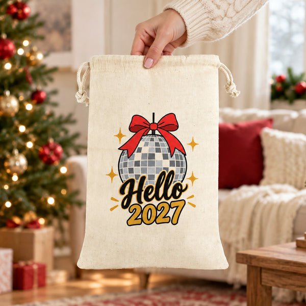 Hand in a cream knit sweater holding a natural drawstring bag with a Hello 2027 disco ball print by its drawstring top in a festive living room, showing the real size