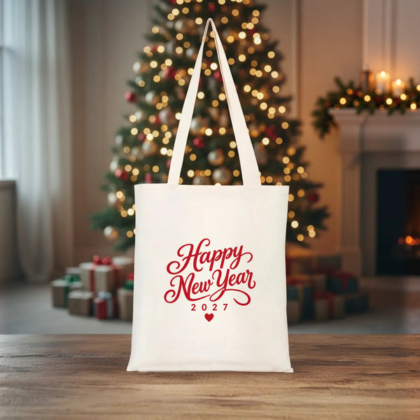Natural cotton gabardine tote bag with a red Happy New Year 2027 script print standing on a wooden table in front of a lit Christmas tree