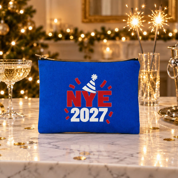 Royal blue clutch bag with an NYE 2027 party hat print standing on a marble bar with champagne glasses, sparklers and gold confetti on New Year's Eve