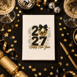 Natural canvas notebook with a 20/27 Happy New Year gold disco ball and bow print on a black New Year's Eve party table with champagne and gold confetti