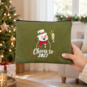 Hand in a cream knit sweater holding an olive green clutch with a Cheers to 2027 snowman print in a festive living room, showing the real size