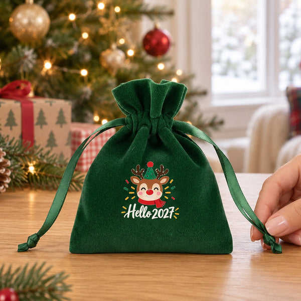 Green velvet drawstring pouch printed with a Hello 2027 reindeer design on a table, a hand in a cream knit sleeve holding its ribbon end to show the real small size