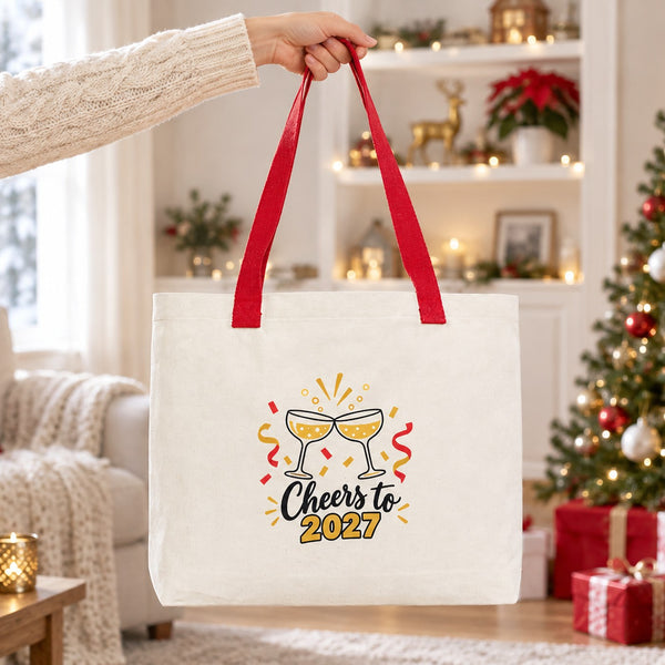 Hand lifting a large canvas tote bag with red knitted straps and a Cheers to 2027 champagne print in a bright Christmas living room