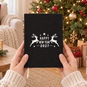 Hands holding a black canvas notebook with an ivory Happy New Year 2027 leaping reindeer print in front of a Christmas tree, showing the real size