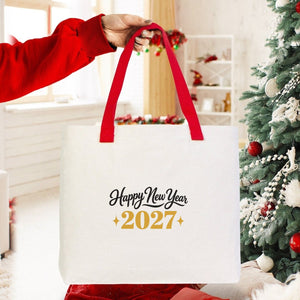 Canvas tote bag with red knitted handles and a Happy New Year 2027 gold print held up beside a Christmas tree