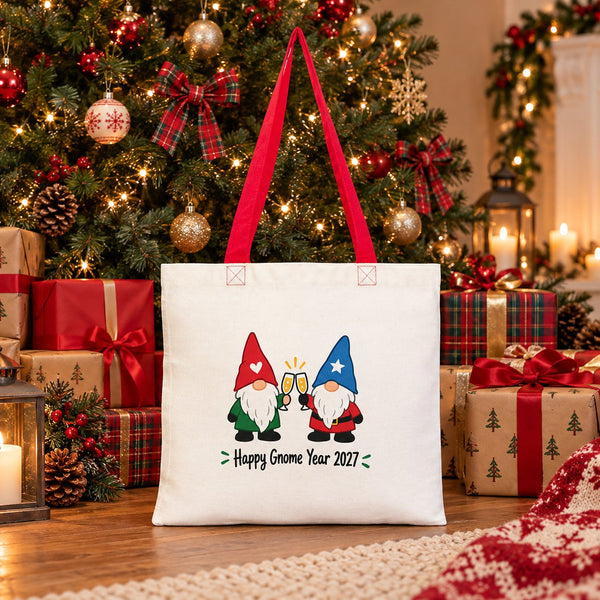 Cotton tote bag with red handles and a Happy Gnome Year 2027 print standing under a Christmas tree among wrapped gifts