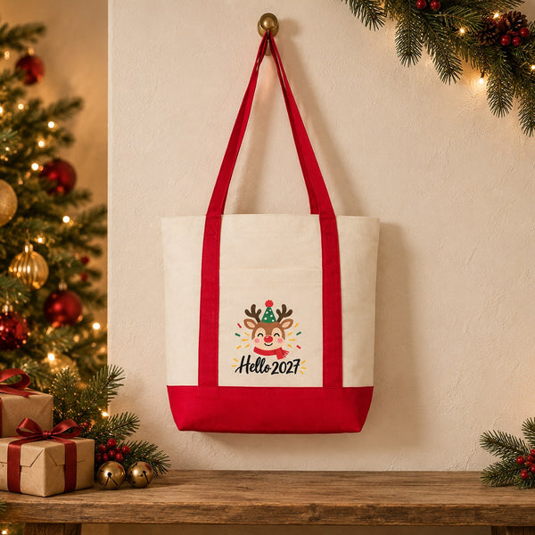 Canvas tote bag with red straps and a reindeer Hello 2027 print hanging on a brass hook beside a Christmas tree