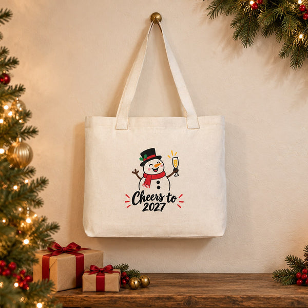 Large canvas tote bag with beige knitted straps and a snowman Cheers to 2027 print hanging on a brass hook beside a Christmas tree