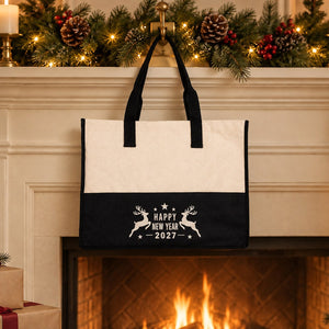 Black canvas tote bag with a Happy New Year 2027 leaping reindeer print hanging from a brass hook on a white fireplace mantel with a garland, candles and a warm fire