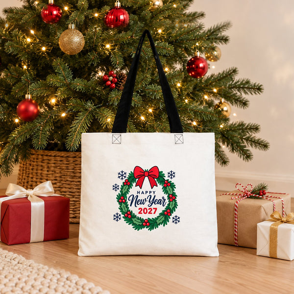 Cotton tote bag with black handles and a holly wreath Happy New Year 2027 print standing by a Christmas tree with three gifts