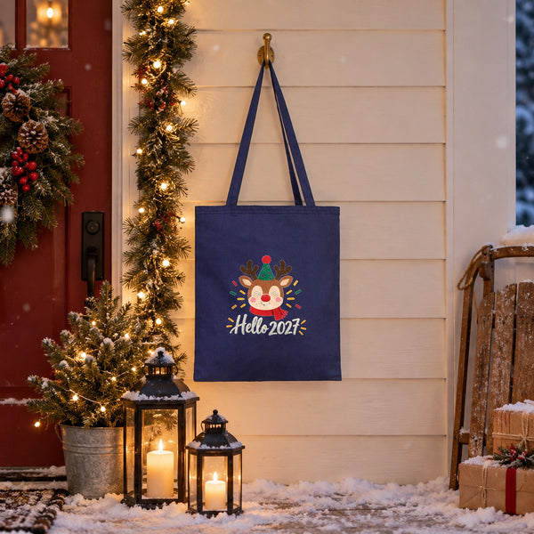 Navy blue cotton tote bag with a Hello 2027 reindeer print hanging by a snowy front door with a Christmas wreath at dusk