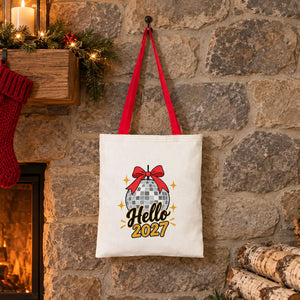 Red handle cotton tote bag with a Hello 2027 disco ball print hanging on a stone wall beside a lit Christmas fireplace