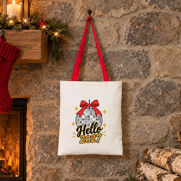 Red handle cotton tote bag with a Hello 2027 disco ball print hanging on a stone wall beside a lit Christmas fireplace