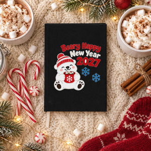 Black canvas notebook with a Beary Happy New Year 2027 polar bear print on a cream knit blanket with hot cocoa, marshmallows and candy canes