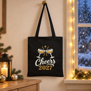 Black cotton gabardine tote bag with an ivory and gold Cheers 2027 champagne print hanging by a snowy window