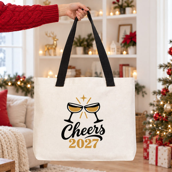 Hand lifting a large canvas tote bag with black knitted straps and a Cheers 2027 champagne print in a bright Christmas living room