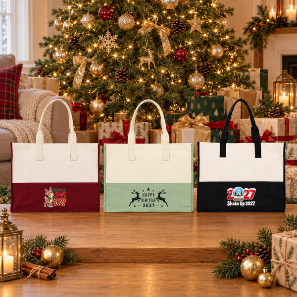 Burgundy, sage green and black canvas tote bags with Merry and Bright reindeer, Happy New Year 2027 reindeer and Shake Up 2027 prints on a table in front of a decorated Christmas tree and presents