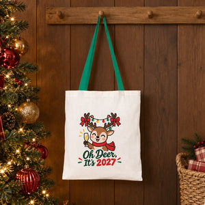 Green handle cotton tote bag with an Oh Deer, It's 2027 reindeer print hanging on a wooden peg rail beside a decorated Christmas tree