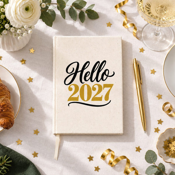 Natural canvas notebook with a Hello 2027 script and gold print on a New Year's Day morning table with coffee, croissant and gold confetti