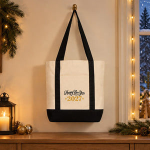 Canvas tote bag with black straps and a Happy New Year 2027 gold print hanging by a snowy window