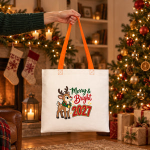 Hand lifting a cotton tote bag with orange handles and a reindeer Merry and Bright 2027 print by a Christmas fireplace in the evening