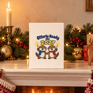 Natural canvas notebook with an Otterly Ready for 2027 otters print standing on a Christmas fireplace mantel with a garland and candles