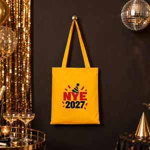 Golden yellow cotton tote bag with an NYE 2027 print hanging at a gold and black New Year's Eve party with champagne