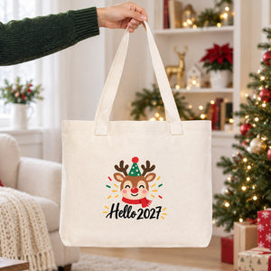 Hand lifting a large canvas tote bag with beige knitted straps and a reindeer Hello 2027 print in a bright Christmas living room