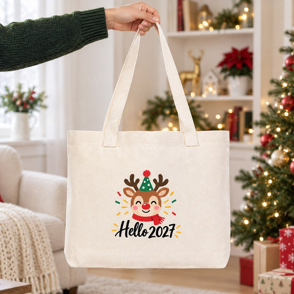 Hand lifting a large canvas tote bag with beige knitted straps and a reindeer Hello 2027 print in a bright Christmas living room