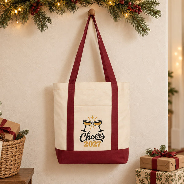 Canvas tote bag with burgundy straps and a Cheers 2027 champagne print hanging on a wooden peg in a Christmas entryway