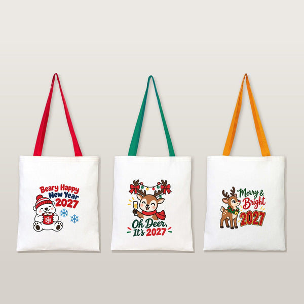Three natural cotton tote bags with red, green and mustard handles and Beary Happy New Year, Oh Deer and Merry and Bright 2027 prints on a plain studio background