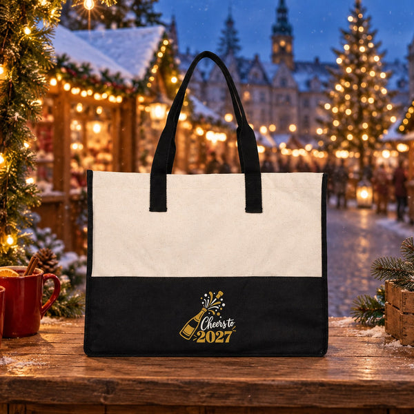 Black canvas tote bag with a Cheers to 2027 champagne print on a wooden Christmas market stall with mulled wine and lanterns