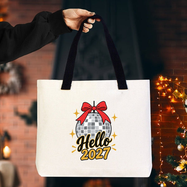 Canvas tote bag with black knitted handles and a Hello 2027 disco ball print held up in a cosy room with fairy lights