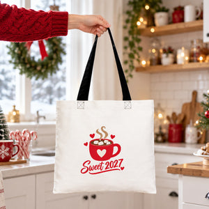Hand lifting a cotton tote bag with black handles and a Sweet 2027 hot cocoa print in a bright Christmas kitchen