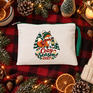 Cream clutch with a green zipper and a Cozy Season 2027 fox print on a buffalo plaid blanket with a lantern, pine cones and cider
