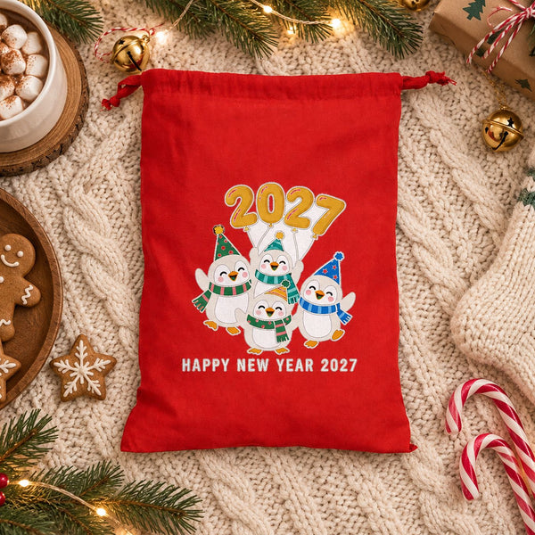 Red drawstring bag with a Happy New Year 2027 penguins print on a cream knit blanket with hot cocoa, gingerbread cookies, candy canes and a knit stocking