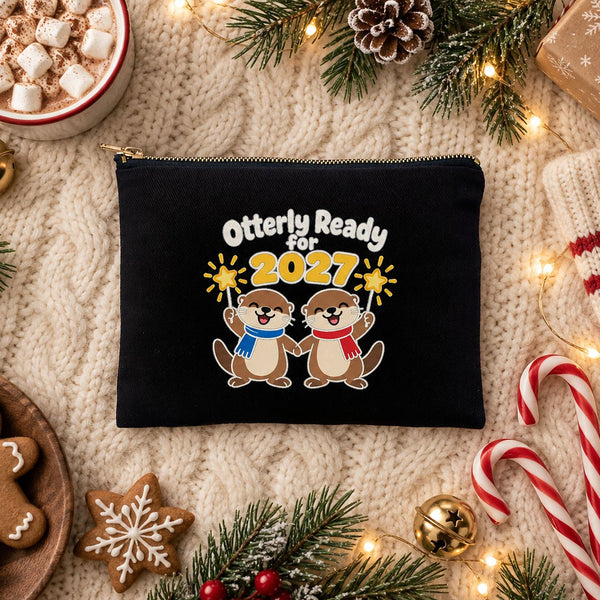 Black clutch bag with an Otterly Ready for 2027 otters print on a cream knit blanket with hot cocoa, a striped stocking, candy canes and gingerbread cookies