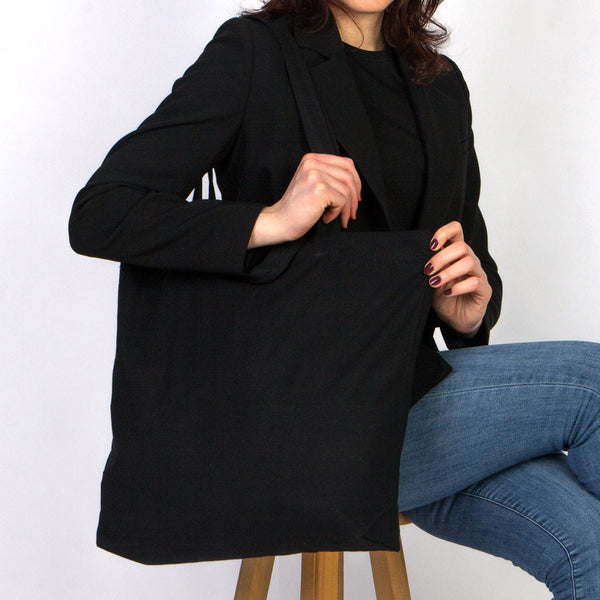 Person wearing a black blazer over a black top and blue jeans, sitting on a wooden stool against a white background.